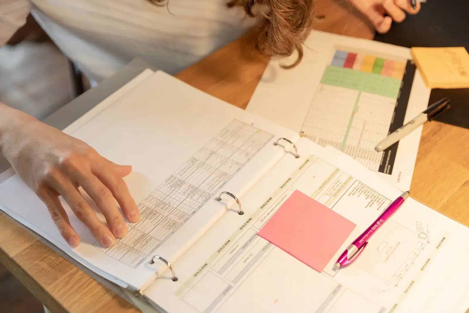 A hand resting on an open ring binder of tabulated pages with sticky notes and pens on a desk